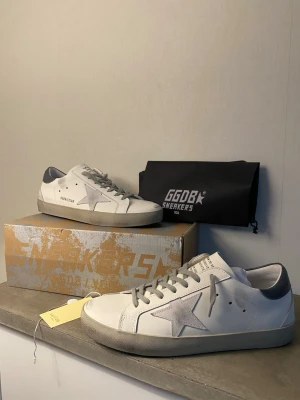 Vita Golden Goose sneakers med grå detaljer - Säljer ett par vita Golden Goose sneakers med grå mockadetaljer och stjärna på sidan. Skorna har en låg silhuett, grå hälkappa i läder och klassisk snörning. Snygg och stilren design som passar till det mesta.