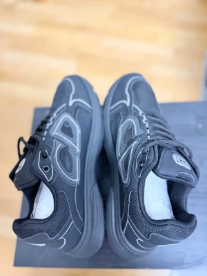 Svarta sneakers med vita detaljer - Snygga svarta sneakers  med vita och grå detaljer. Skorna har en chunky sula, meshpaneler och reflekterande linjer längs sidorna. Klassisk snörning och bekväm vadderad plös. Perfekta för dig som gillar streetwear och exklusiv stil.