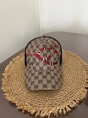 Gucci keps med orm och GG-mönster - Snygg keps från Gucci med klassiskt GG-mönster i beige och brunt. Framtill pryds kepsen av en röd, vit och svart orm. Baksidan är i svart mesh för extra ventilation. Perfekt accessoar för dig som vill sticka ut med lyxig streetstyle.