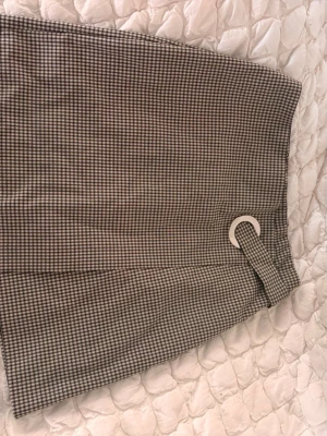 Rutig kortkjol med bältesdetalj - Säljer en snygg rutig kortkjol i beige och svart med en cool bältesdetalj framtill. Kjolen har en rak passform och är perfekt för dig som gillar klassisk men trendig stil. Materialet känns som bomull eller polyester och mönstret är smått rutigt.