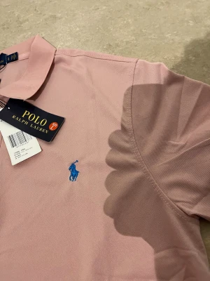 Rosa pikétröja från Polo Ralph Lauren - Säljer en klassisk rosa pikétröja från Polo Ralph Lauren med blå broderad logga på bröstet. Tröjan har korta ärmar, krage och knappar vid halsen. Perfekt för en stilren och avslappnad look.