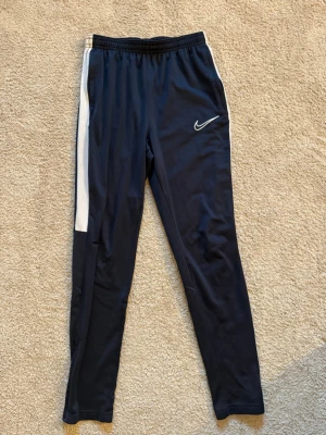 Mörkblåa Nike Dri-FIT träningsbyxor  - Mörkblåa Nike Dri-FIT träningsbyxor med vit sidopanel och elastisk midja. Byxorna har fickor och dragkedjor vid änden av benen. Perfekta för träning eller sportaktiviteter.