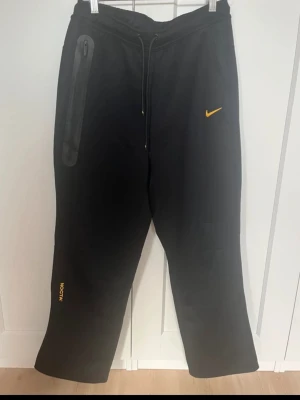 Svarta mjukisbyxor från Nike - Svarta mjukisbyxor från Nike med gul logga och gul text på vänster ben. Byxorna har dragsko i midjan och en dragkedjeförsedd ficka på höger sida. Avslappnad passform och mjukt material, perfekta för chill dagar.