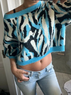 Blå och svart mönstrad cropped tröja - Säljer en stickad cropped tröja med blå, svart och vit zebramönstrad design. Tröjan har bred halsringning som kan bäras off-shoulder och ribbade kanter. Perfekt för dig som vill ha en statement piece i garderoben.