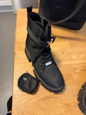 Steve Madden skor - Säljer ett par svarta kängor med grov sula och snörning. Kängorna har ett brett spänne över skaftet och är tillverkade i ett slitstarkt tygmaterial. Perfekta för dig som gillar en tuff och modern stil.