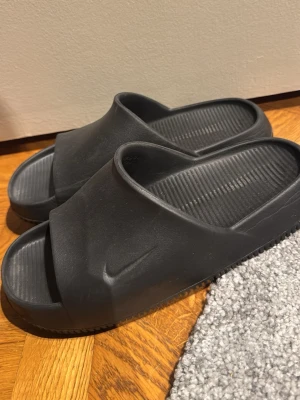 Svarta slides från Nike - Svarta tofflor från Nike i en stilren och enkel design. Tillverkade i ett mjukt och flexibelt gummimaterial med räfflad sula för extra grepp. Perfekta att glida i hemma eller till och från träningen. Diskret Nike-logga på sidan.