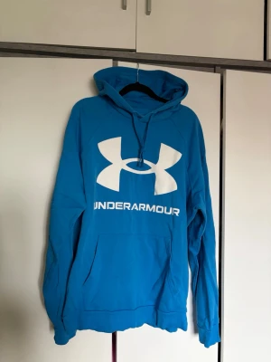 Blå hoodie från Under Armour - Snygg blå hoodie från Under Armour med stor vit logga och text på bröstet. 