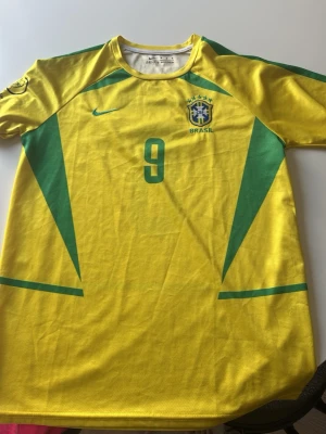 Brasilien fotbollströja Ronaldo #9 - Gul Brasilien fotbollströja från Nike med gröna detaljer och nummer 9 på bröstet och ryggen. RONALDO står tryckt på ryggen. Tröjan har korta ärmar och är tillverkad i ett lätt och ventilerande material, perfekt för fotbollsträning eller match.