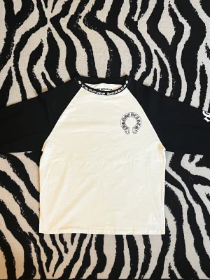 Svartvit långärmad topp Chrome Hearts S - Säljer en svartvit långärmad topp från Chrome Hearts med kontrastärmar och tryck både fram och bak. Toppen har logga på bröstet, stort tryck på ryggen och textdetaljer på ärmarna samt runt halsen. Perfekt för dig som gillar streetwear och grafiska prints.
