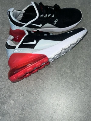 Nike Air Max 270 svart/röd/vit, Helt nya ej använda  - Nike Air Max 270 sneakers med svart ovandel, vita och grå detaljer samt en stor röd Air Max-sula. Skorna har en sportig design med tydlig logga på sidan och en luftig meshstruktur upptill. Perfekta för dig som gillar streetstyle och bekväma sneakers.