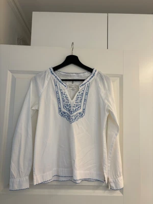Vit blus Lexington - Vit blus från Lexington med blå broderade detaljer längs v-ringningen och ärmsluten. Blusen har lång ärm och är gjord i ett tunt, mjukt tyg.