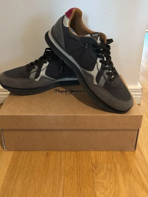 Grå sneakers från Pepe Jeans - Snygga sneakers från Pepe Jeans i olika nyanser av grått med svarta och vita detaljer samt en vinröd hälflik. Insidan är i läder och i hög kvalité! Skorna är i bra skick och skolåda medföljer i köp. Skorna är även snörade på ett fint sätt men snörningen kan jag även ta bort vid köp. Ny pris: 1350. Storlek 43!