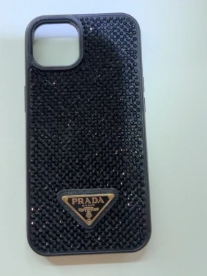 Svart glittrigt mobilskal från Prada - Snyggt svart mobilskal från Prada täckt med små glittrande stenar och en ikonisk metallbricka med loggan längst ner. Skalets form är anpassad för en iPhone 13 och ger en lyxig känsla med sin blingiga yta. Perfekt för dig som vill ha något extra till mobilen.