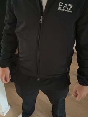 Svart vindjacka från Emporio Armani - Snygg svart vindjacka från Armani EA7 med dragkedja framtill och logga på bröstet. Jackan har en sportig look, hög krage och sidofickor med dragkedja.