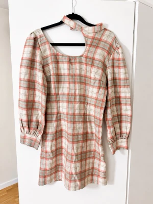 Rutig kortklänning med unik hals - Trendig kortklänning med rutigt mönster i beige, rosa och grå toner. Klänningen har långa ärmar, markerade muddar och en unik halsringning med asymmetrisk detalj. Perfekt för dig som vill sticka ut med en cool och modern look.