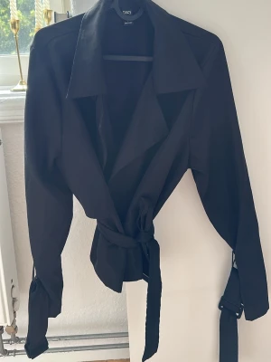 Svart trenchcoat med knytskärp - Stilren svart trenchcoat med bred krage och knytskärp i midjan. Jackan har långa ärmar med spänndetaljer vid handlederna och en klassisk, rak passform. Perfekt för en trendig och tidlös look.