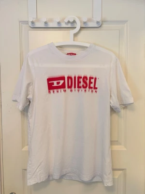 Vit t-shirt från Diesel med röd logga - Väldigt snygg Diesel T-shirt i väldigt bra skick! 0 fläckar eller skavanker.  📐 Längd: 66 cm Axelbredd: 43 cm Öppning runt midja: 51 cm Halsöppning: 20 cm  Hör av dig vid frågor och funderingar!🤝  Fraktar så fort jag kan!📦