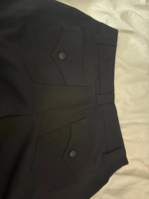 Svarta byxor Gina Tricot XXS - Svarta byxor från Gina Tricot med två bakfickor med lock och knapp. Byxorna har hällor för skärp och stängs med hake och knapp framtill. Snygg och enkel design som passar till många olika stilar.