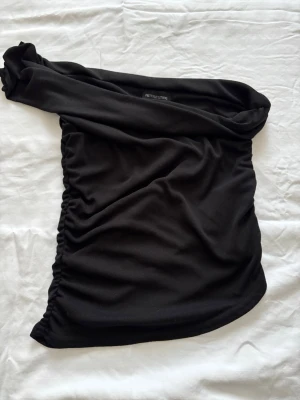Svart oneshoulder topp från PrettyLittleThing - Säljer en svart oneshoulder topp från PrettyLittleThing. Toppen är tight och har en snygg rynkad detalj på sidan. Perfekt för dig som vill ha en stilren och trendig look med en axel bar. Materialet känns mjukt och stretchigt.
