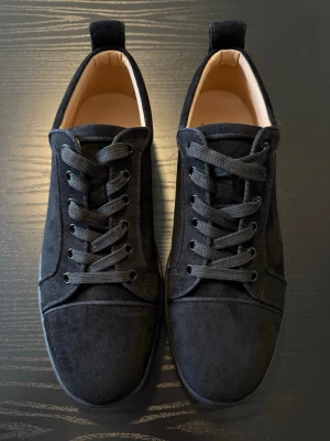 Svarta loubutin sneakers i mocka-look - Stilrena svarta sneakers med mocka-liknande material och svarta skosnören. Klassisk låg modell med rund tå och diskret design. Passar perfekt till jeans eller streetwear.
