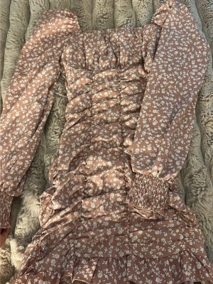 Blommig kort klänning med volanger - Supersöt kort klänning i beige med vitt blommönster. Klänningen har långa puffärmar med smock vid ärmslut, volangdetaljer och rynkningar framtill. Perfekt för sommarens alla tillfällen och ger en romantisk vibe.