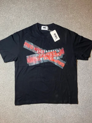 Masion margiela t shirt - Svart t-shirt från MM6 Maison Margiela med unikt grafiskt tryck i vitt och rött framtill. Klassisk passform och rund halsringning. Tillverkad i mjuk bomull som känns skön mot huden. Perfekt för dig som gillar statement-plagg och streetwear vibes.