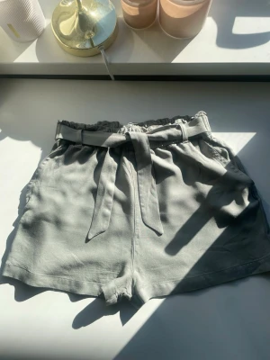 Grå shorts med knyt från KappAhl - Snygga grå shorts från KappAhl med hög midja och knytband i midjan. Shortsen har en avslappnad passform och är perfekta för varma dagar. Tillverkade i ett mjukt material som känns skönt mot huden.