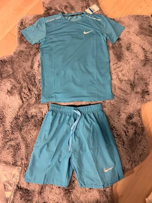Nike ljusblått träningsset miler shorts - Nike träningsset i ljusblått med t-shirt och shorts. T-shirten har rund hals och vita detaljer vid axlarna samt vit Nike-logga på bröstet. Shortsen har resår och dragsko i midjan samt vit Nike-logga på benet. Perfekt för träning eller sport.