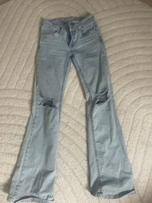 Ljusblå bootcut jeans med slitningar - Säljer ett par ljusblå bootcut jeans med slitna hål på båda knäna och klassiska fem fickor. Jeansen har en utsvängd passform nedtill och är gjorda i denim. Snygga detaljer på bakfickan med broderad söm.