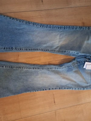 Ljusblå bootcut jeans med nitar - Säljer ett par ljusblå bootcut jeans med snygga detaljer i form av nitar på bakfickorna. Jeansen har klassisk femficksmodell och är tillverkade i denim med en lätt tvättad look. Perfekta för dig som gillar en avslappnad men trendig stil.