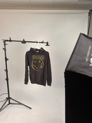 Svart Nirvana hoodie med tryck - Säljer en svart hoodie med huva och magficka, prydd med det klassiska Nirvana-logotypen och smileytrycket i gult på bröstet. Perfekt för dig som gillar bandmerch och vill ha en avslappnad, cool stil. Dragsko i huvan och ribbade muddar.