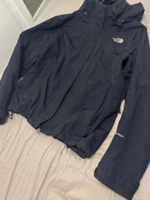 Svart regnjacka från The North Face - Snygg svart regnjacka från The North Face med HyVent-teknologi. Jackan har huva med dragsko, hel dragkedja framtill och två sidofickor med dragkedja. Perfekt för blöta dagar och outdoor-äventyr.