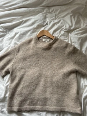 Beige stickad t-shirt i ullmix - Mysig beige stickad t-shirt med korta ärmar och rund hals. Tröjan har en avslappnad passform och ribbade kanter vid ärmslut och nederkant. Perfekt för lager på lager och ger en soft look till din outfit.