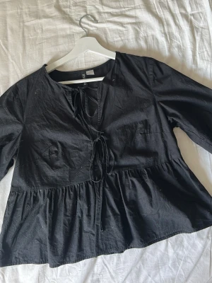 Svart blus med knytning fram - Svart blus i luftig bomull med v-ringning och dekorativa knytband framtill. Modellen har en avslappnad passform och trekvartsärm. Perfekt till jeans eller kjol för en trendig look.