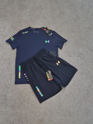 Under Armour  - Snyggt träningsset från Under Armour med t-shirt och shorts i mörkblått. Båda plaggen har färgglada logotyper och tryckdetaljer. Materialet är lätt och snabbtorkande, perfekt för träning eller sportaktiviteter. T-shirten har rund hals och shortsen har resår i midjan.