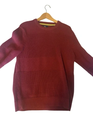 Vinröd stickad tröja från RLS - Snygg vinröd stickad tröja från RLS med rund halsringning och bred ribbstickad kant nedtill och vid ärmslut. Tröjan har en strukturerad stickning i olika partier som ger en cool look. Perfekt för dig som gillar stilrena och bekväma plagg.