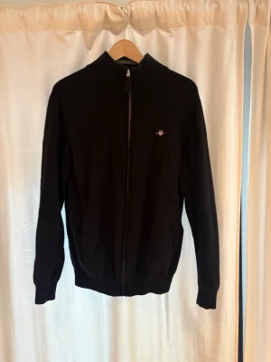 Svart ziptröja med liten Gant logga - Svart långärmad Gant ziptröja med hög krage och liten broderad logga på bröstet. Tröjan har ribbade muddar vid ärmslut, är stretchig och nederkant. Perfekt för lager-på-lager och enkel att matcha med olika outfits.