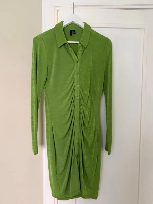 Grön skjortklänning från Vero Moda - Snygg grön skjortklänning från Vero Moda med långa ärmar och knappar framtill. Klänningen har en cool rynkad detalj längs magen och är tillverkad i ett mjukt, stretchigt material. Perfekt för dig som vill sticka ut med färg och stil!