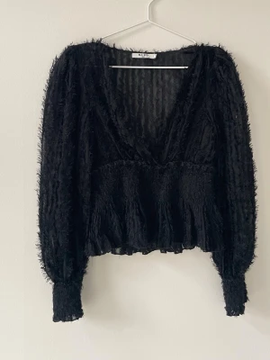 Svart v-ringad blus från NA-KD - Snygg svart blus från NA-KD med v-ringning och lång ärm. Blusen har en transparent look med struktur och fransiga detaljer, samt smock vid ärmslut och midja. Perfekt för dig som vill ha en trendig och unik topp.