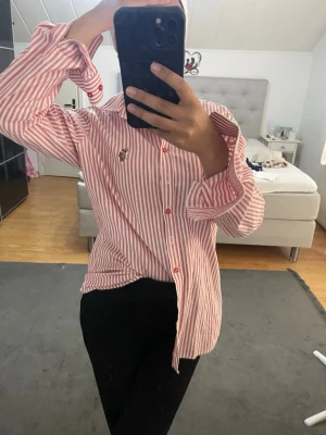 Rödvit randig skjorta med brodyr polo  - Säljer en röd och vit randig skjorta med långa ärmar och klassisk krage. Skjortan har röda knappar och en liten broderad detalj på bröstet. Perfekt för en trendig och avslappnad look. Storlek L men passar som S-M