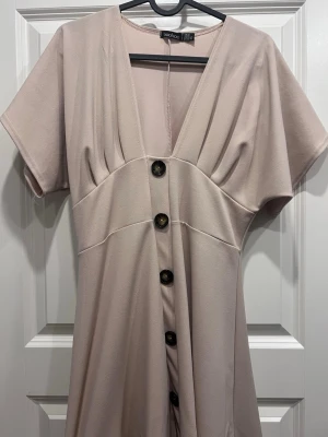 Beige kortklänning med knappar från  Boohoo - Snygg beige kortklänning från Boohoo med v-ringning, stora dekorativa knappar framtill och korta ärmar. Klänningen har en lös passform och är tillverkad i ett mjukt syntetmaterial. Perfekt för dig som gillar enkel men trendig stil.
