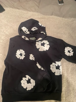 Svart hoodie med vita blommor - Cool svart hoodie med stora vita blommönster tryckta över hela tröjan, inklusive huvan. Hoodien har klassisk passform, ribbade muddar och en mjuk känsla. Perfekt för dig som vill sticka ut med en unik och trendig stil.