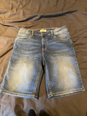 Blå jeansshorts från Lindex - Snygga blå jeansshorts från Lindex med klassisk femficksdesign och gul kontrastsöm. Shortsen har normal passform och stängs med knapp och dragkedja. Perfekta för varma dagar och enkla att matcha med olika toppar.