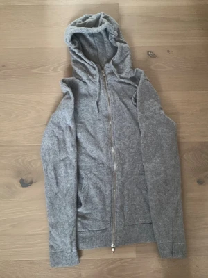 Kashmir Zip Hoodie NNO7 - Mysig grå hoodie från NN07 med hel dragkedja framtill och justerbar huva med snören. Tillverkad i mjukt material och har ribbade muddar vid ärmslut och nederkant. Perfekt för en avslappnad och stilren look. Passar dig som är runt 165-175cm