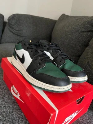 Nike Air Jordan 1 Low svart/grön - Snygga Nike Air Jordan 1 Low sneakers i svart, grönt och vitt läder. Klassisk siluett med svart swoosh på sidan och gul Jumpman-logga på plösen. Perfekta för dig som gillar streetwear och vill ha en ikonisk sneaker i garderoben.