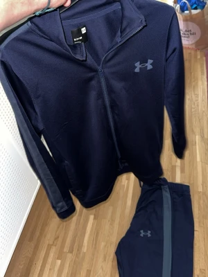Marinblått träningsset från Under Armour - Snyggt marinblått träningsset från Under Armour med ziptröja och matchande byxor. Setet har diskreta loggor på både tröja och byxa samt en grå rand längs byxbenet. Perfekt för gymmet eller löprundan. Tillverkat i ett mjukt och stretchigt funktionsmaterial.