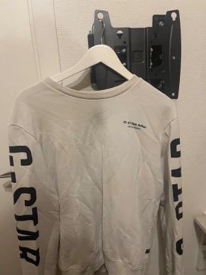 Vit sweatshirt från G-Star Raw - Vit sweatshirt från G-Star Raw med tryckt logga på bröstet och stora svarta bokstäver längs båda ärmarna. Tröjan har rund halsringning och ribbade muddar vid ärmslut och nederkant. Perfekt för en avslappnad streetwear-look.