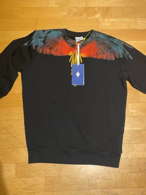 Svart tröja Marcelo Burlon med vingar - Säljer en svart sweatshirt från Marcelo Burlon County of Milan med ikoniskt färgglatt vingtryck över axlar och bröst i rött, orange, blått och gult. Rund halsringning och ribbade muddar. Perfekt statement-plagg för dig som vill sticka ut.