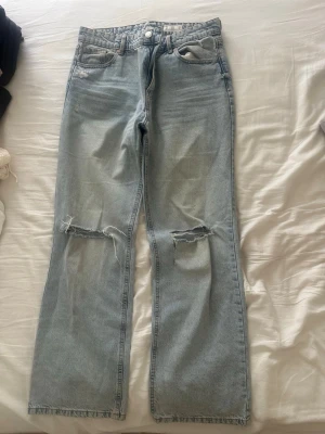 Ljusblå slitna bootcut jeans - Säljer ett par ljusblå bootcut jeans med slitningar och hål på båda knäna. Jeansen har klassisk femficksmodell, knappgylf och är tillverkade i denim. Perfekta för en avslappnad och trendig look.