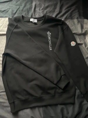 Svart Moncler sweatshirt - Snygg svart sweatshirt från Moncler med vit logotyptryck på bröstet och klassisk Moncler-patch på ärmen. Tröjan har rund halsringning, ribbade muddar och en clean, avslappnad passform. Perfekt för en stilren och modern look.